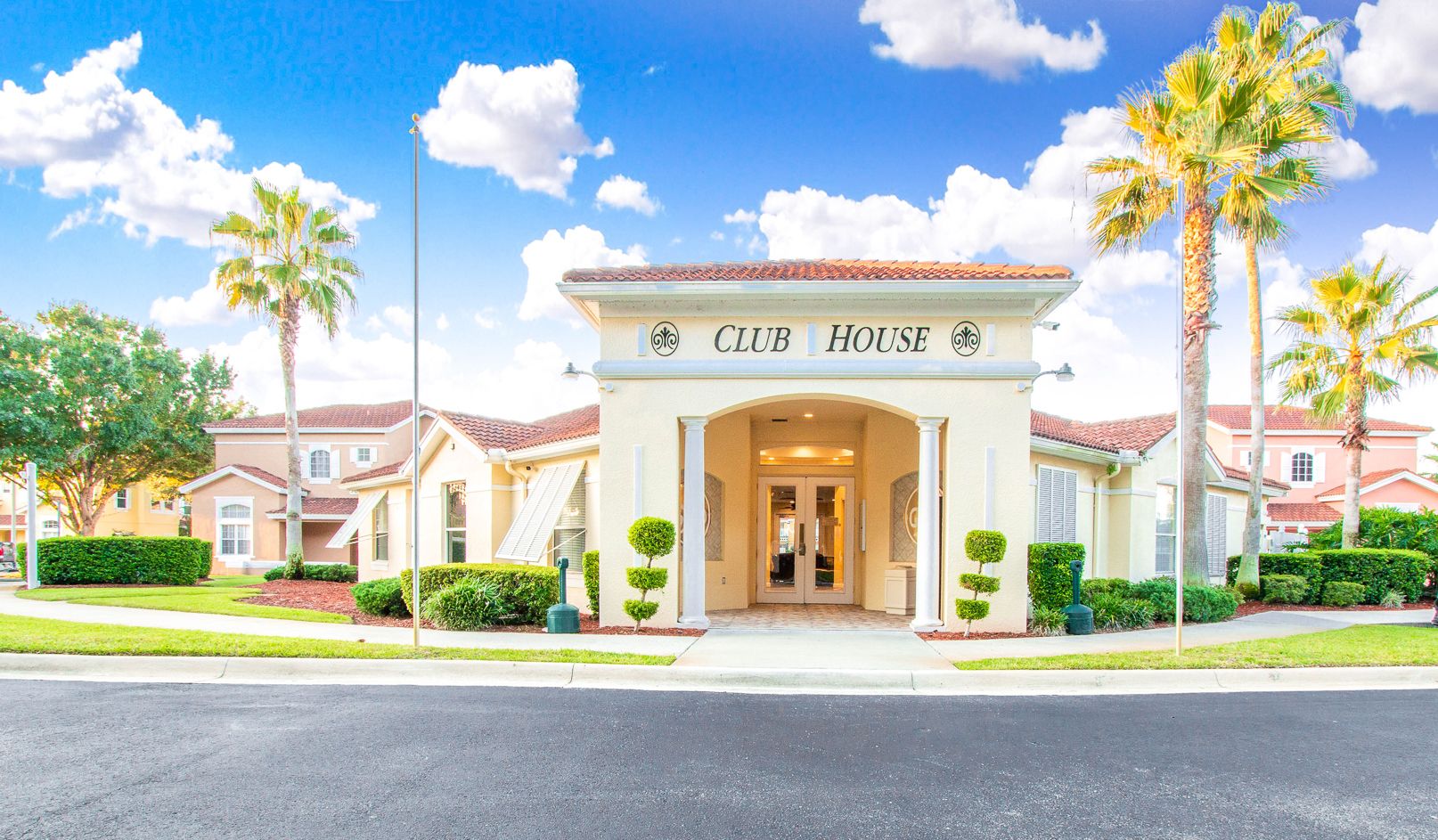 A cream-colored building with a tiled roof, arched entrance, and columns, surrounded by palm trees and landscaping.